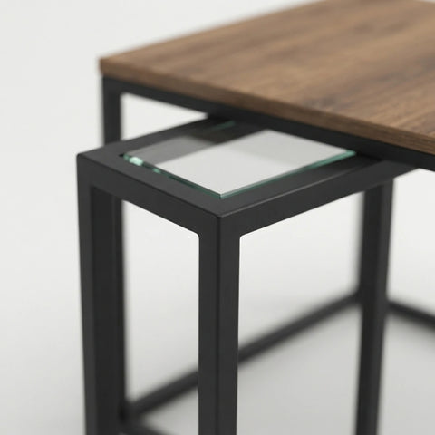 Close-up of a nesting table set with a medium brown wood grain top and matte black metal frame, featuring a clear glass insert on the front table.