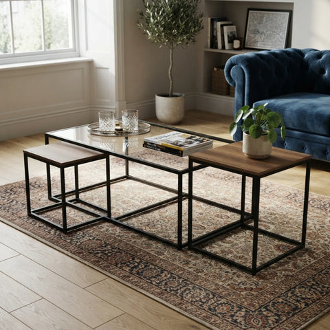 A nesting coffee table set in a bright living room. The set includes a rectangular glass-top coffee table with a wood top and two matching square nesting side tables with wood tops and black metal frames, all arranged on a patterned area rug.