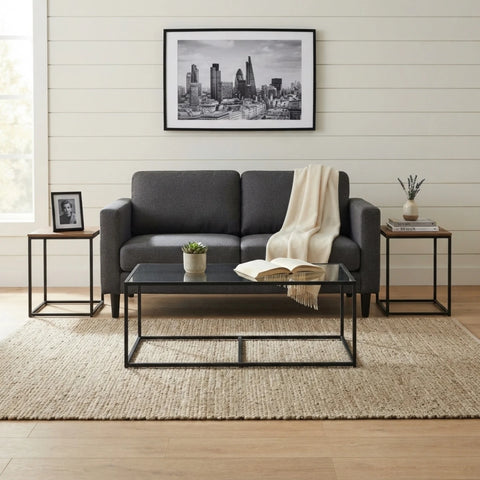 A modern living room setup featuring a dark gray two-seater sofa with a cream throw blanket, a black glass-top coffee table, and two matching wood-and-black side tables on a woven beige rug against a white shiplap wall.