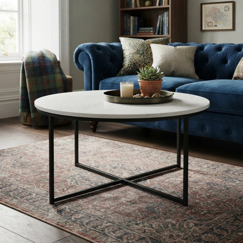 A round coffee table with a white stone top and black metal legs, styled in a living room setting with a blue sofa, patterned rug, and decorative tray on top.