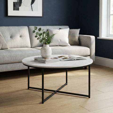 A round coffee table with a white marble-look top and black metal legs, styled in a living room with a light gray sofa, dark blue wall, and wood flooring.
