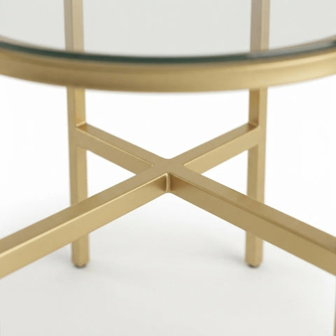 Close-up of a round glass-top table with a polished gold metal base featuring an X-shaped leg support and slim vertical legs.