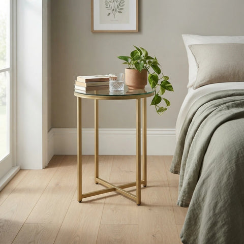 A round glass-top side table with a gold metal frame and legs, styled in a bedroom next to a bed with a potted plant and books on top.