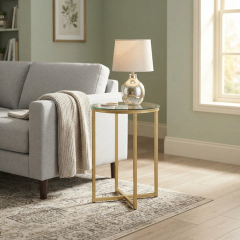 A modern round glass-top end table with a gold metal frame, styled in a living room next to a gray sofa with a cream throw blanket and a matching table lamp.