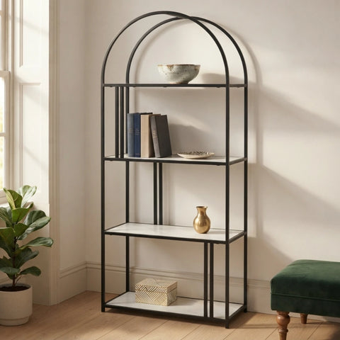 A black metal shelving unit with five white shelves stands in a bright room next to a window, holding books, a bowl, a small vase, and a box. A potted plant is on the left and a green velvet stool is on the right.