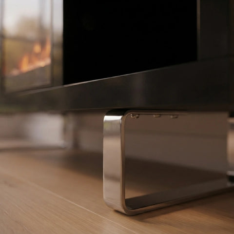 A close-up of a modern flat-screen TV resting on a polished metallic stand over a light wood floor, with a blurred fireplace in the background.