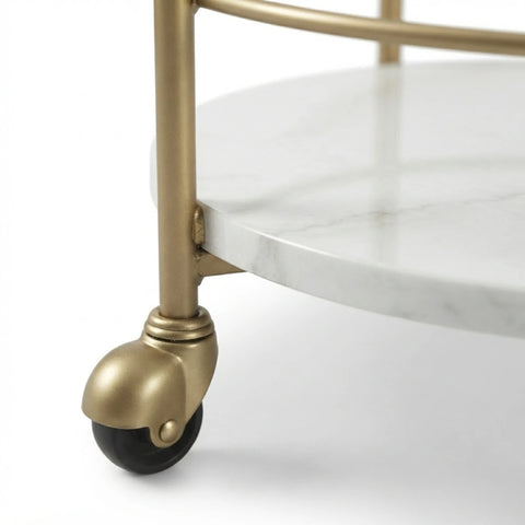 Close-up of a bar cart or rolling side table with a white marble round tray, brushed gold metal frame, and black caster wheel on a white background.