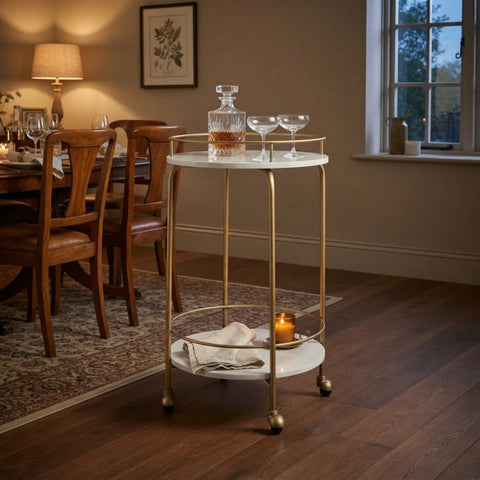 A round bar cart with a white marble-look top and bottom shelf and a gold metal frame, styled in a warm dining room setting with wine glasses, a decanter, and candles.