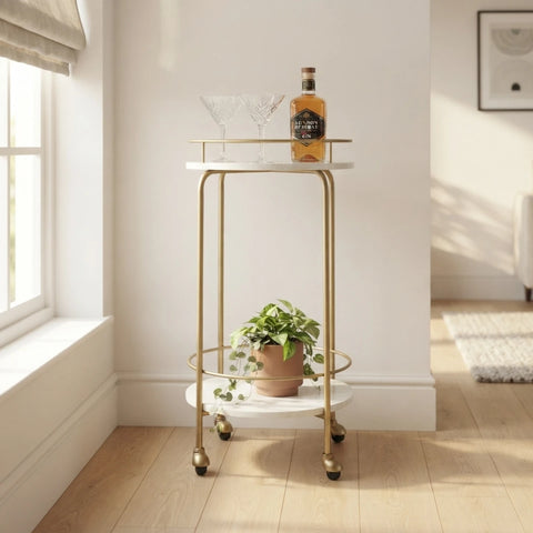 A two-tier bar cart with a white marble-look tray and gold metal frame, styled with a bottle, glasses, and a potted plant in a bright room.