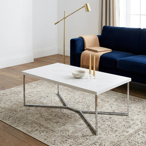 A modern rectangular coffee table with a white marble-look top and a chrome X-shaped metal base, styled in a living room setting on a patterned area rug.