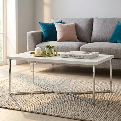 A modern rectangular coffee table with a white top and chrome metal legs, styled in a bright living room with a gray sofa, textured rug, and decorative items.